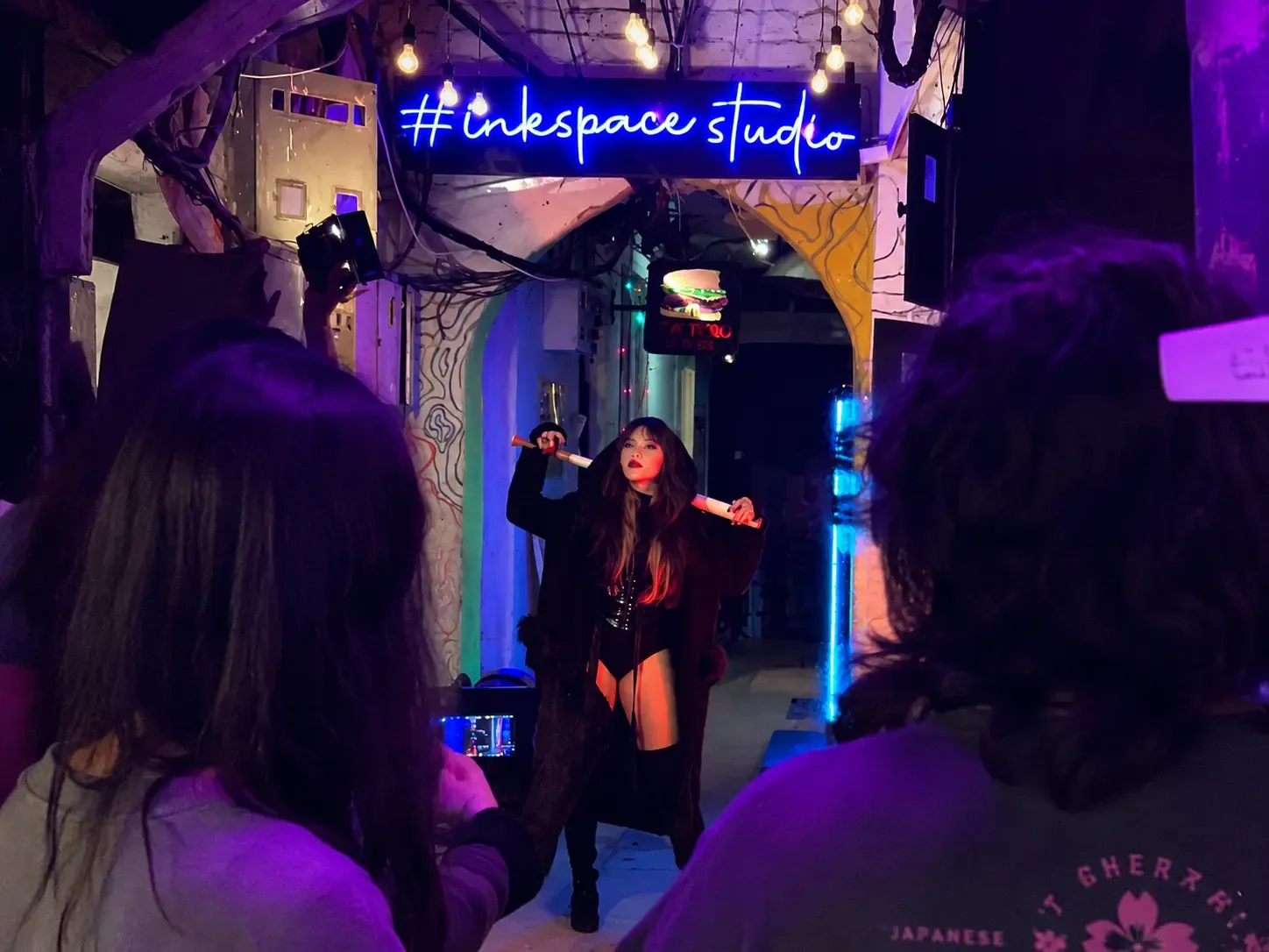 Shoot under the #inkspacestudio neon sign A performer posing under the blue neon #inkspacestudio sign in Space Gully while onlookers film on their phones.