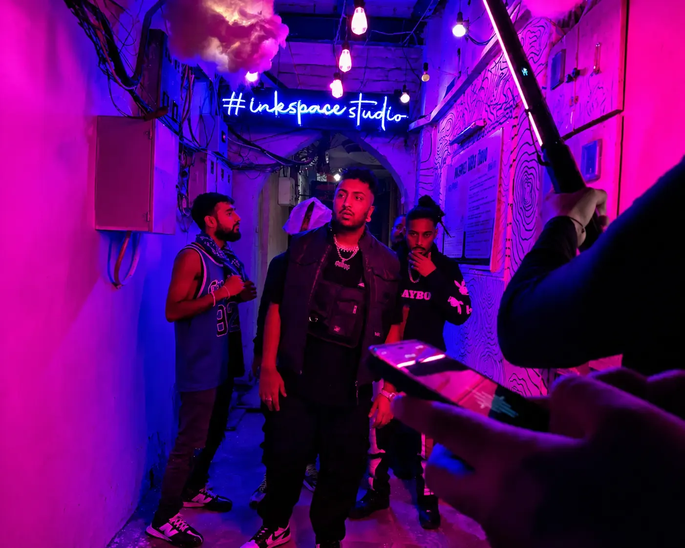 Space Gully neon lane during a shoot A group of musicians standing in the pink and blue neon-lit Space Gully lane under the #inkspacestudio sign during a video shoot.