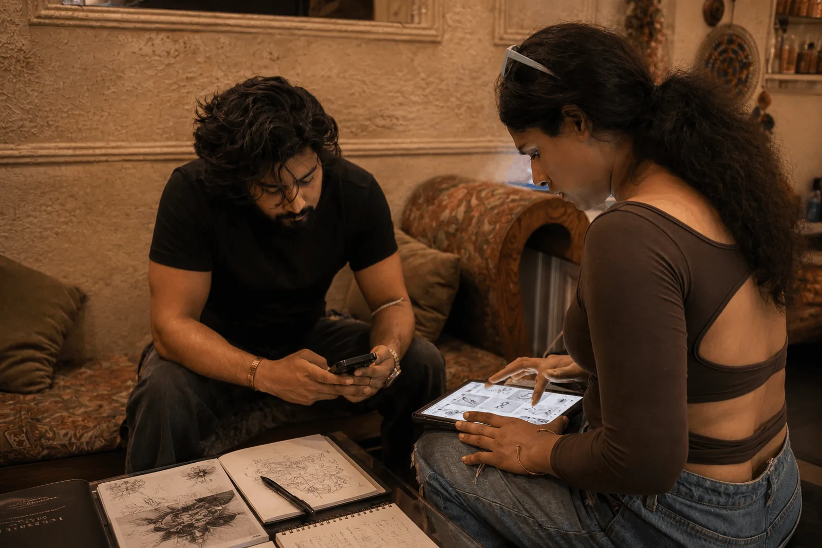 Custom tattoo consultation at Inkspace A tattoo artist and a client sitting on a studio sofa, the client scrolling reference sketches on a tablet while open sketchbooks of hand-drawn designs lie on the table in front of them.