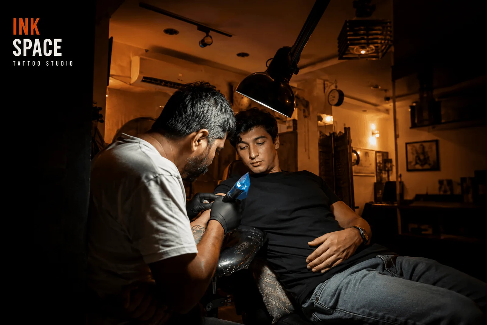 Tattooing in progress at Inkspace A tattoo artist leaning in with a machine to work on a reclining client's forearm under an angled studio lamp, surrounded by framed artwork and warm studio lighting.