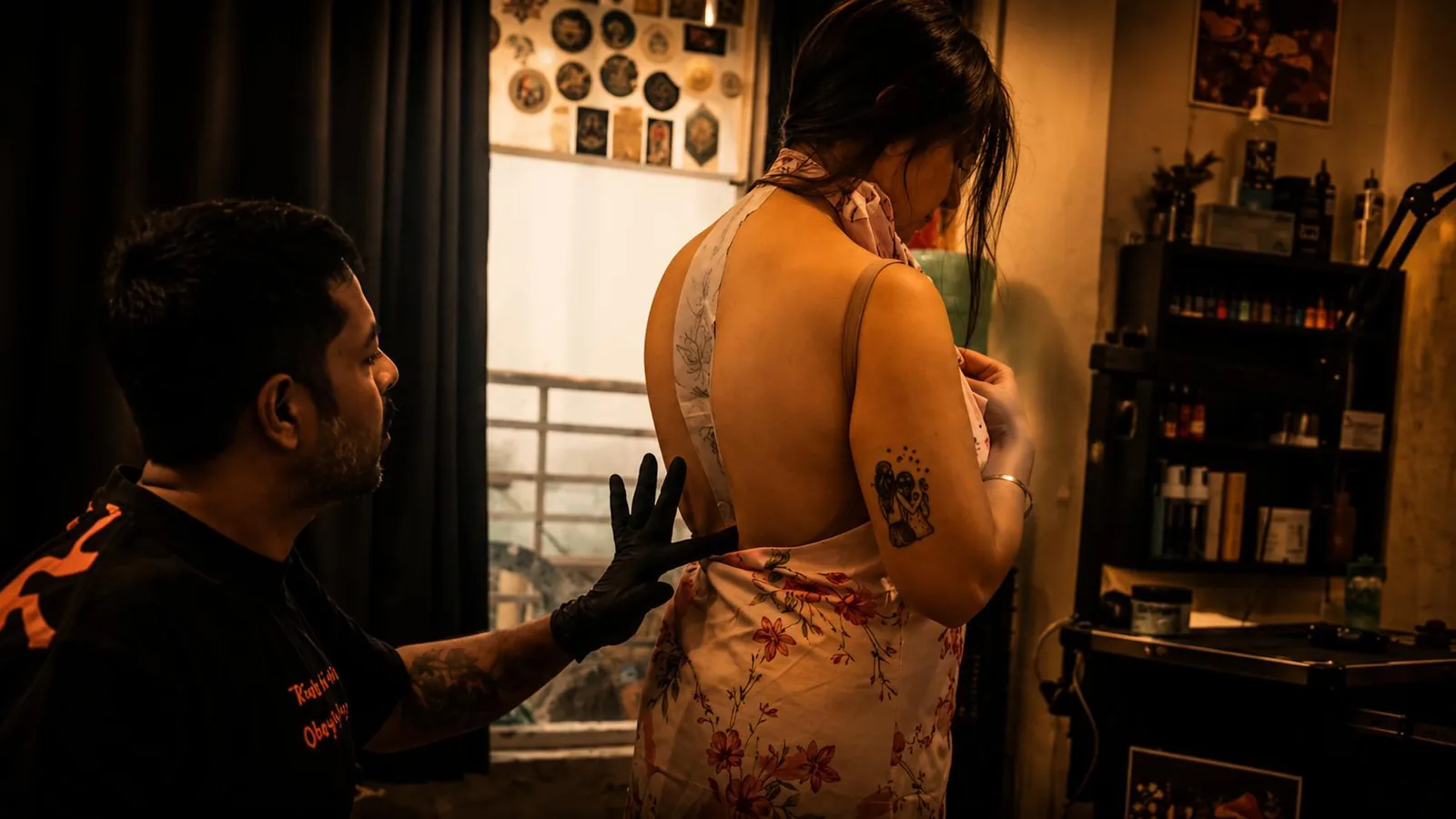 Planning the right tattoo placement A gloved tattoo artist gesturing towards a standing client's upper back as they discuss where a new piece will sit, with framed tattoo artwork on the studio walls behind them.